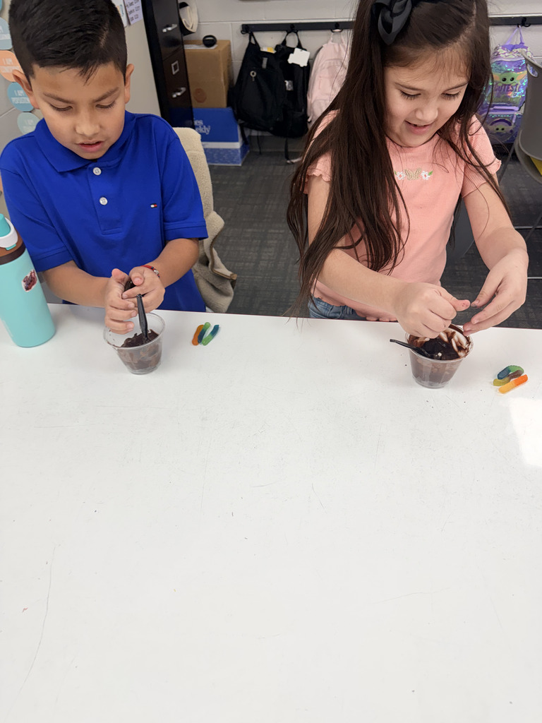 wo elementary students work together at a table, carefully adding gummy worms to clear cups filled with chocolate "dirt" pudding