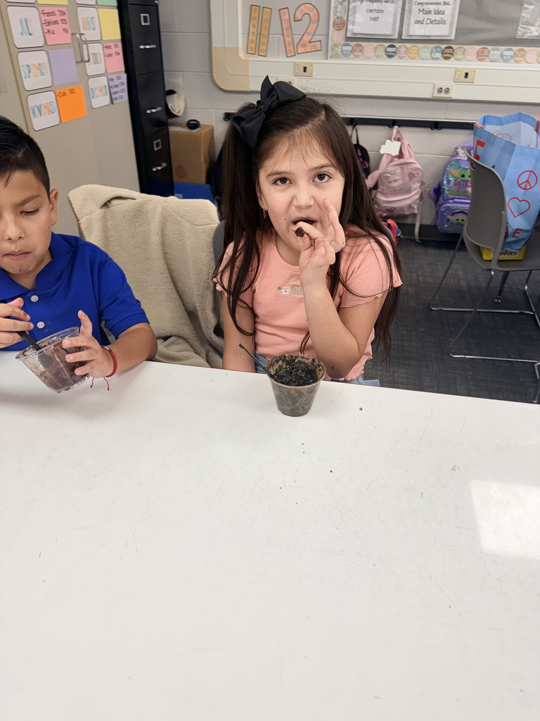 A young elementary student sitting at a classroom desk takes a bite of a homemade chocolate pudding treat topped with crushed cookies from a small clear cup.