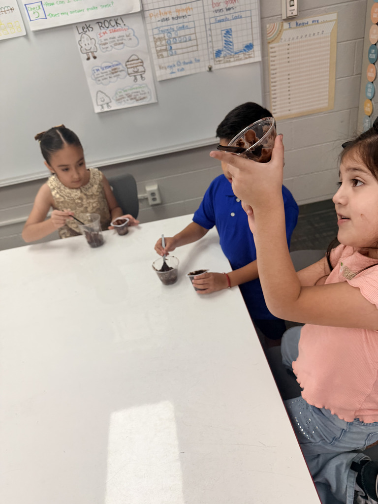 A young girl holds up her clear cup of chocolate dirt dessert in a classroom while other students sit nearby working on their treats.