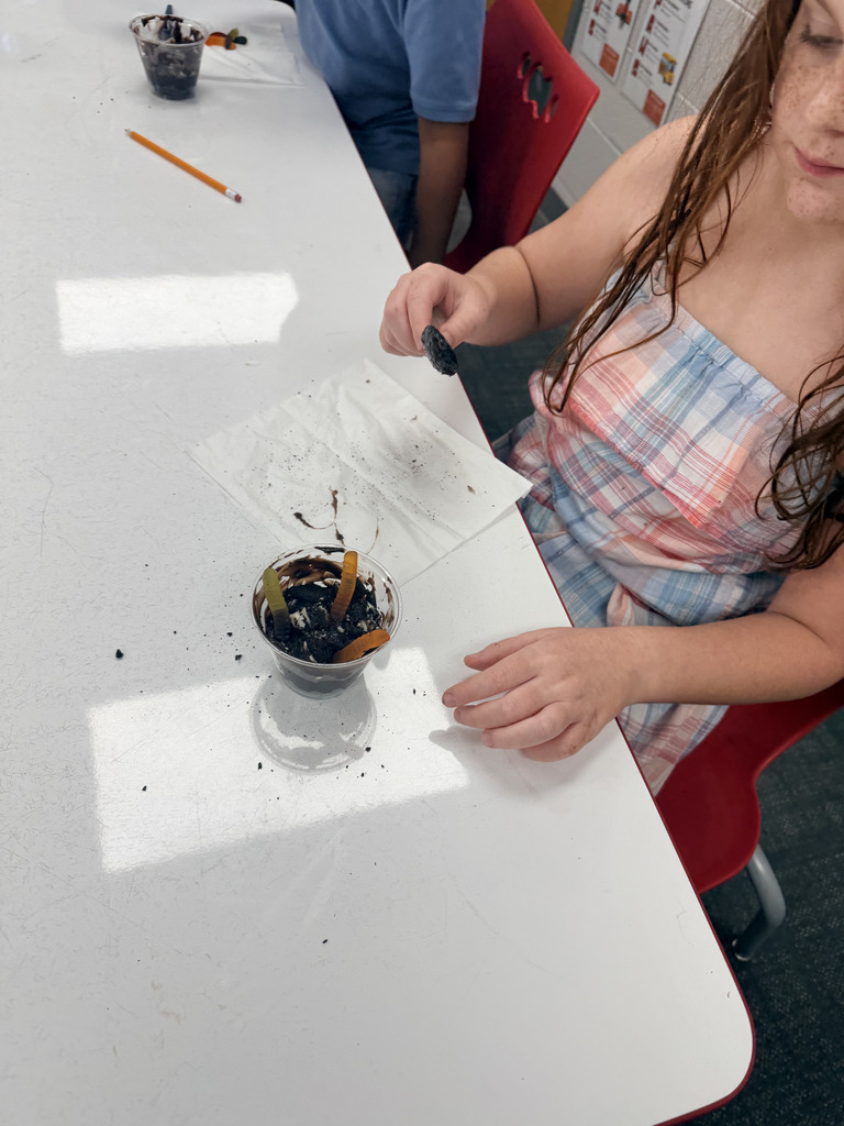 Close-up view of a student placing a chocolate cookie into a clear plastic cup filled with chocolate pudding and gummy worms.