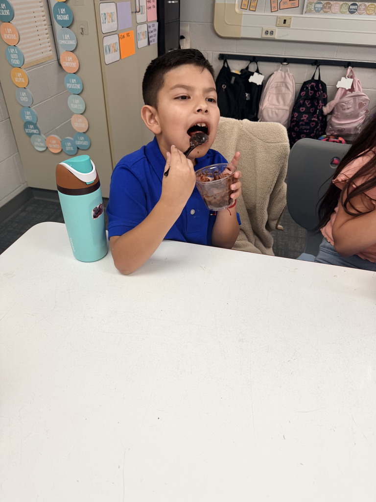 A young boy in a blue polo shirt eats a spoonful of chocolate dirt pudding at his classroom table.