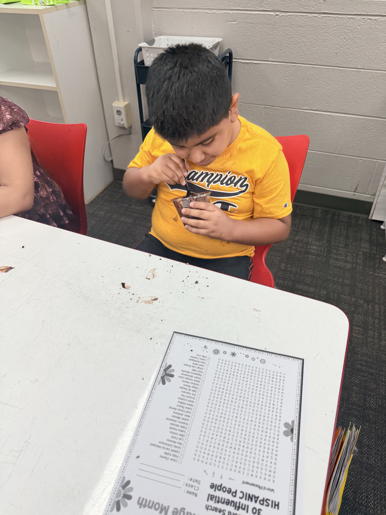 A student wearing a yellow shirt looks down attentively while eating his cup of chocolate dessert at a classroom desk, next to a Hispanic Heritage Month word search worksheet.