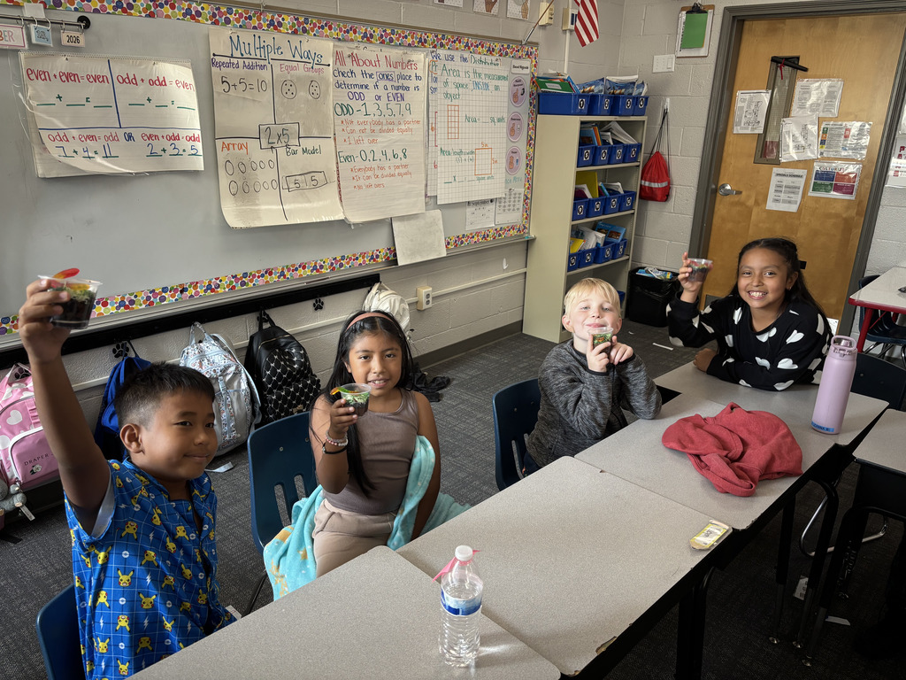 Four elementary students seated at desks in front of a whiteboard hold up their finished clear cups of chocolate dirt pudding and smile for the camera.