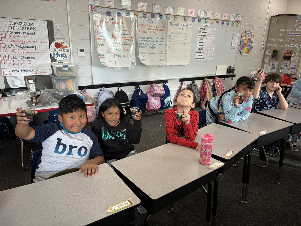 Five young students sit at their classroom desks holding up their clear cups filled with chocolate pudding, crushed cookies, and gummy worms.