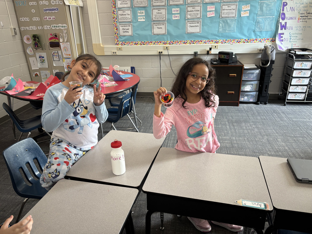 wo smiling elementary students hold up their completed cups of chocolate dirt pudding while sitting at their classroom desks in pajamas.