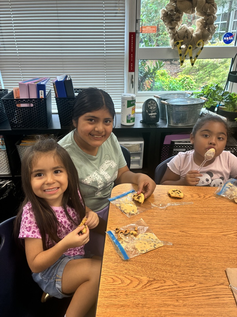 A young girl in a pink patterned top smiles at the camera while holding a cookie, sitting at a classroom desk next to an older student in a light green shirt who smiles broadly. Across from them, a third student in a panda shirt eats with a plastic spoon. Several cookies, plastic snack baggies, and spoons are laid out on the wooden table, with window blinds, plants, and classroom storage bins visible in the background.