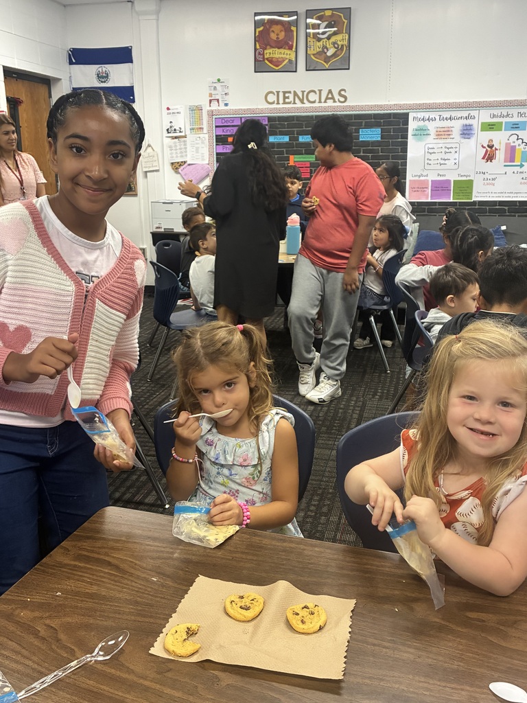 A young girl with pigtails in a baseball-patterned dress smiles at the camera while sitting at a wooden table next to another young girl in a floral top who is eating with a white plastic spoon. Standing beside them, an older girl wearing a pink and white heart-patterned cardigan holds a small baggie and spoon while looking toward the camera. Three chocolate chip cookies rest on a napkin on the table. In the busy classroom background, other students move around near science displays labeled "CIENCIAS," measurement charts, and Hogwarts house crest posters.