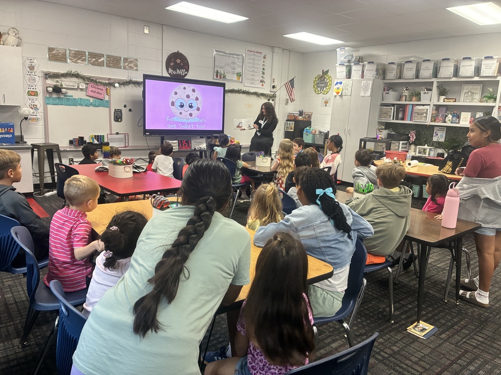 A group of young elementary students sitting at flexible seating tables in a bright classroom. They are facing a monitor screen showing an illustrated chocolate chip cookie with the phrase "Reading Buddies are Sweet too." A teacher sits near the display at the front of the room.