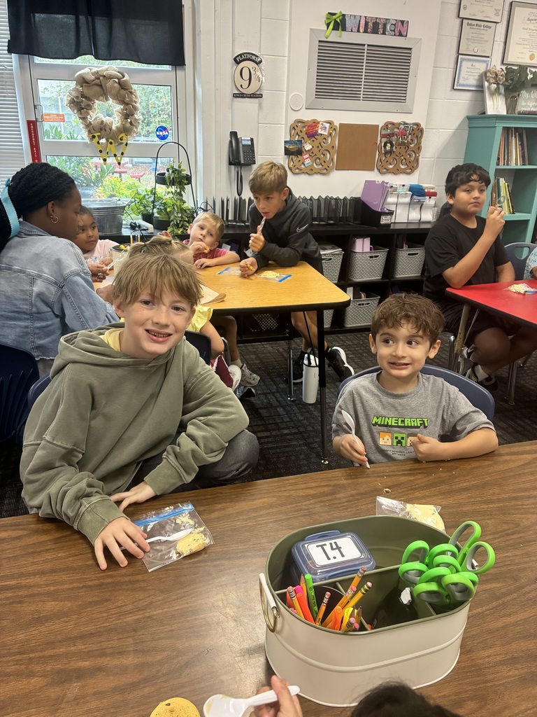 A smiling student in an olive green hoodie sits at a wooden classroom desk next to a younger boy wearing a grey Minecraft t-shirt. On the table in front of them sits a white supply caddy labeled "T.4" filled with scissors and pencils, alongside plastic snack baggies and spoons. In the background, other students are seated at nearby desks throughout the classroom, which features a "Platform 9 ¾" sign and classroom decor on the wall.