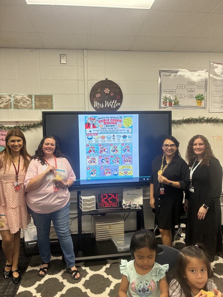 Four women stand smiling in front of a large digital monitor displaying a poster titled "Westie the Wildcat's Edible Cookie Dough Recipe!" in a classroom setting. Two young students sit in the foreground. The classroom wall behind them features a sign reading "Mrs. Witten" alongside instructional diagrams, and a digital clock below the screen reads 12:24.