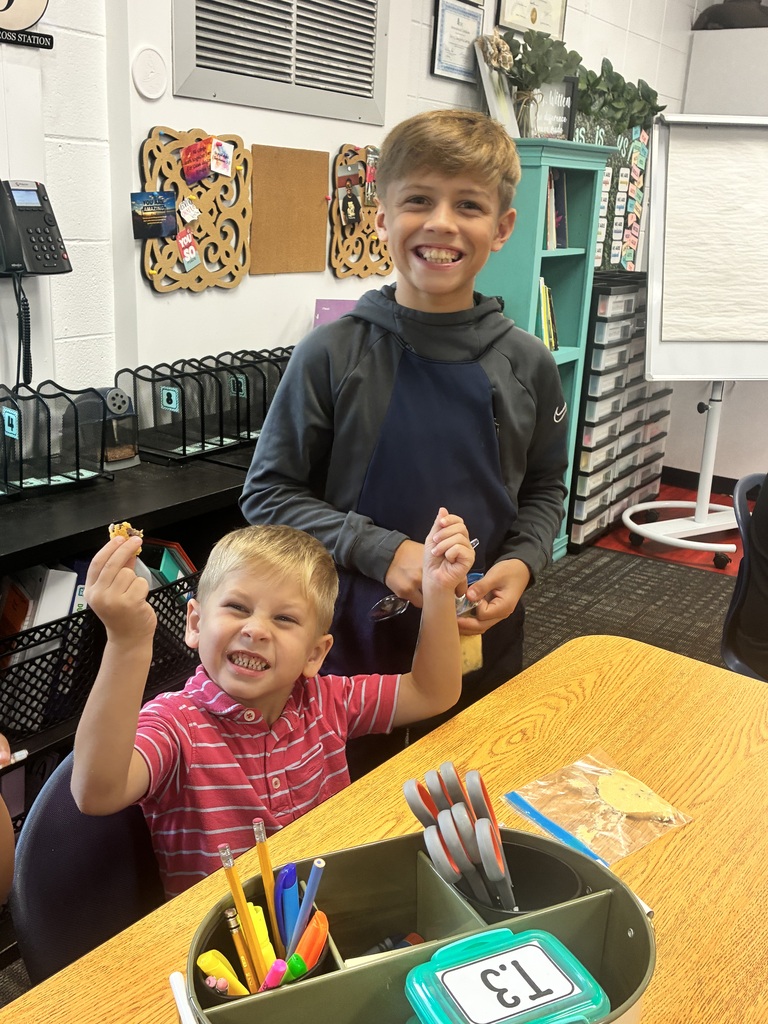 A young boy in a pink striped polo shirt sits at a classroom desk, smiling excitedly while holding up a small treat. Behind him, an older boy in a grey sweatshirt and apron smiles broadly. School supplies sit on the desk in front of them.