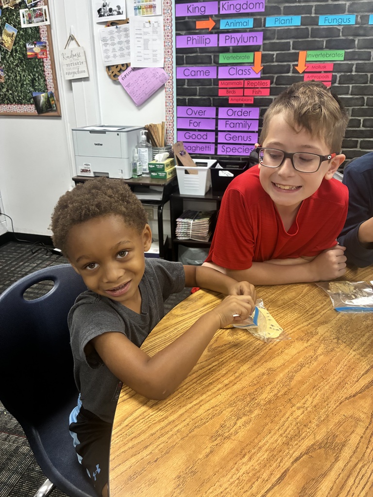 A young boy in a grey shirt smiles at the camera while opening a small clear baggie on a wooden table. Beside him, an older boy wearing glasses and a red shirt smiles as he leans over the table to watch. In the background, a classroom wall features a taxonomy chart outlining biological classification ranks like Kingdom, Phylum, and Class.