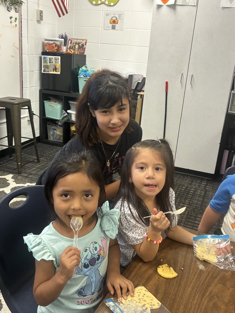 A young girl wearing a light blue Stitch graphic shirt smiles with a clear plastic spoon in her mouth, sitting next to another young girl in a floral top who holds her own spoon while looking at the camera. Leaning in behind them is an older student with dark ponytail hair wearing a dark shirt. Cookies and plastic snack bags rest on the wooden classroom table in front of them, with storage cabinets and shelves visible in the background.