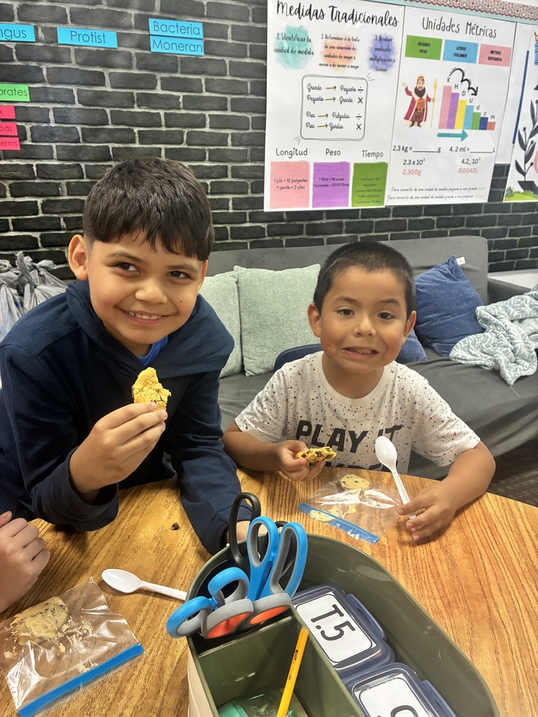 Two young boys sit together at a wooden classroom desk, smiling while enjoying cookies. The boy on the left in a dark blue hooded sweatshirt holds up a bite of his cookie, while the boy on the right in a white patterned shirt holding the text "PLAY" smiles with a cookie piece in hand and a plastic spoon nearby. On the desk in front of them is a supply organizer labelled "T.5" holding scissors and pencils, along with clear plastic snack bags. In the background, posters about "Medidas Tradicionales" and "Unidades Métricas" hang against a faux black-brick wall behind a grey couch.