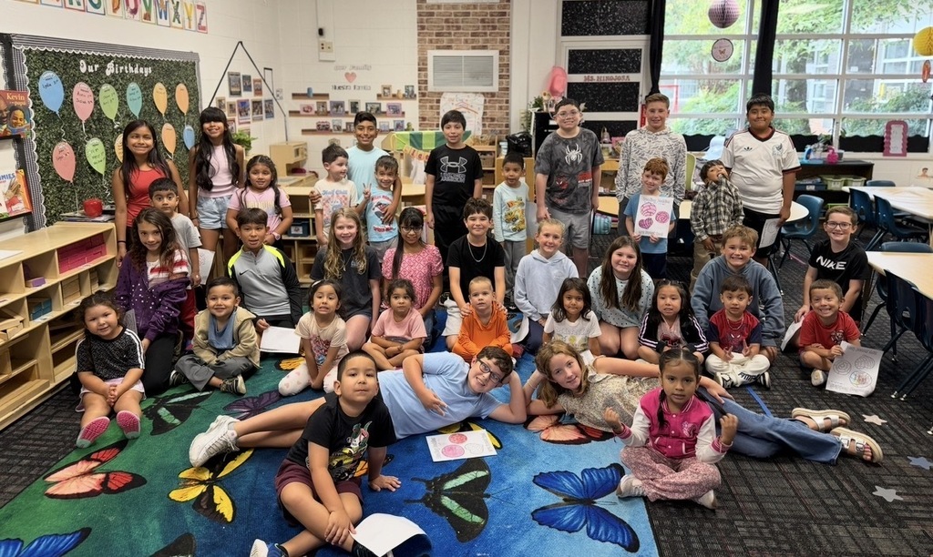 A large group of elementary students poses together for a photo in a brightly decorated classroom. Some children stand along the back, while others sit or lounge comfortably across a colorful butterfly-patterned rug in the foreground. Several students hold up "Reading Buddies" coloring pages, showcasing their completed artwork from the paired activity.