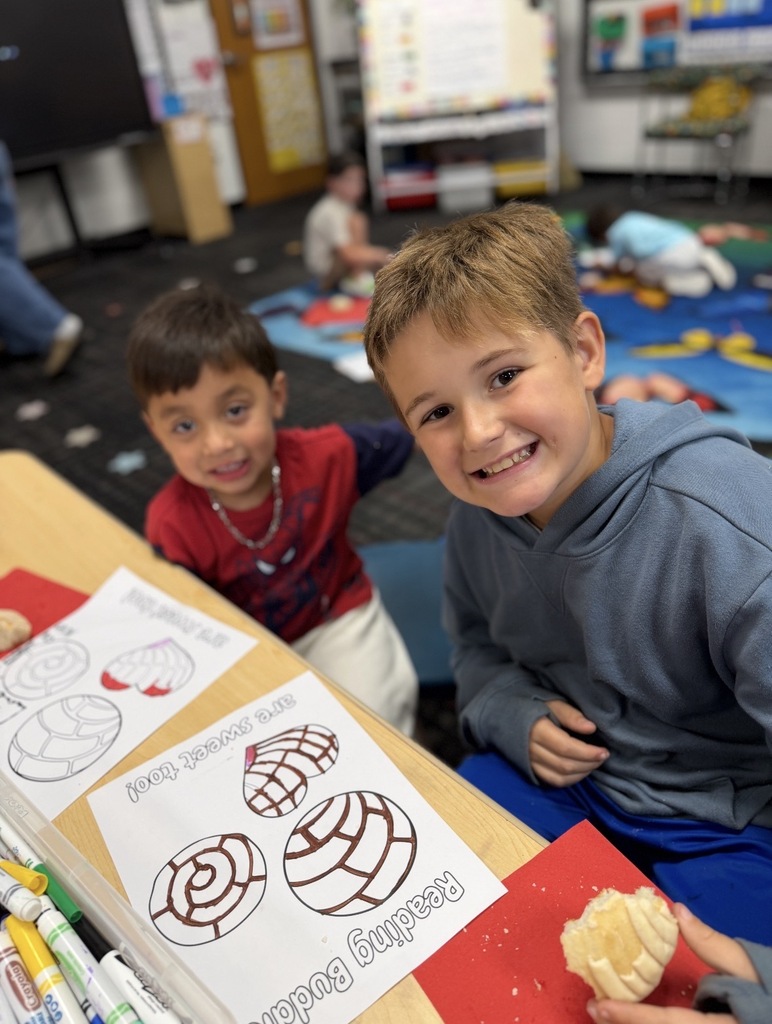 Two young boys smile at the camera while sitting at a classroom table during a "Reading Buddies" activity. The boy on the right in a blue hooded sweatshirt holds a piece of sweet bread (concha), while the boy on the left wears a red graphic long-sleeve shirt. On the desk in front of them are markers and "Reading Buddies are sweet too!" coloring pages featuring conchas.