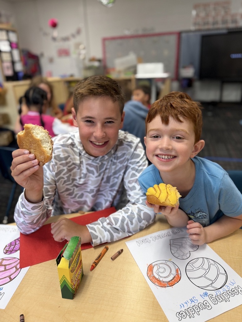 Two smiling boys sit at a classroom desk, holding up pieces of sweet bread (conchas) during a "Reading Buddies" activity. The older boy on the left wears a patterned gray long-sleeve shirt and holds a partially eaten bread piece, while the younger red-haired boy on the right wears a blue T-shirt and holds a piece with yellow icing. Crayons and coloring sheets featuring pan dulce designs are spread across the table in front of them.