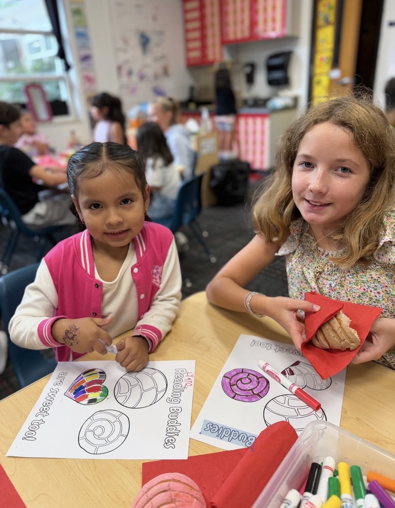 Two young girls smile at a classroom table during a "Reading Buddies" activity. The younger girl on the left wears a pink varsity-style jacket and holds a marker, while the older girl on the right holds a piece of sweet bread (concha) on a red napkin. In front of them are "Reading Buddies are sweet too!" coloring sheets featuring pan dulce illustrations, along with markers and a pink play dough concha.