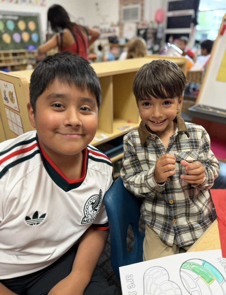 Two smiling young boys pose together during a classroom activity. The older boy on the left wears a white soccer jersey featuring the Mexico national team emblem, while the younger boy on the right wears a plaid button-down shirt. In front of them on the table sits a partially colored "Reading Buddies" worksheet featuring pan dulce designs.