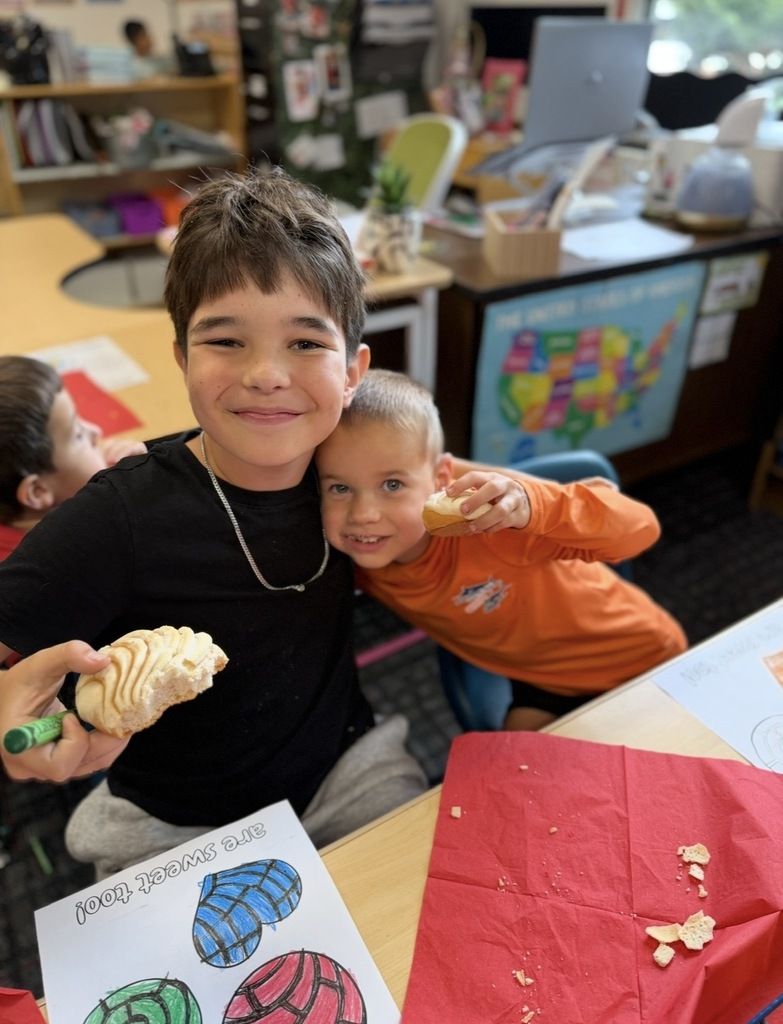 Two smiling young boys pose close together at a classroom desk during a Reading Buddies activity. The boy on the left wears a black shirt, holds a green marker, and shows off a partially eaten piece of sweet bread (concha). The younger boy on the right, wearing an orange long-sleeve shirt, hugs him while holding his own small piece of bread. A colored pan dulce worksheet and a red paper napkin sit on the desk in front of them.