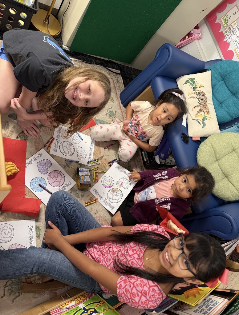 Four students sit together on the rug in a classroom reading corner, surrounded by crayons and "Reading Buddies are sweet too!" coloring sheets featuring pan dulce. The students include an older girl in a pink printed top and glasses sitting in the foreground, two younger girls seated nearby on the floor, and an older girl leaning over from the left, all participating in the activity together.