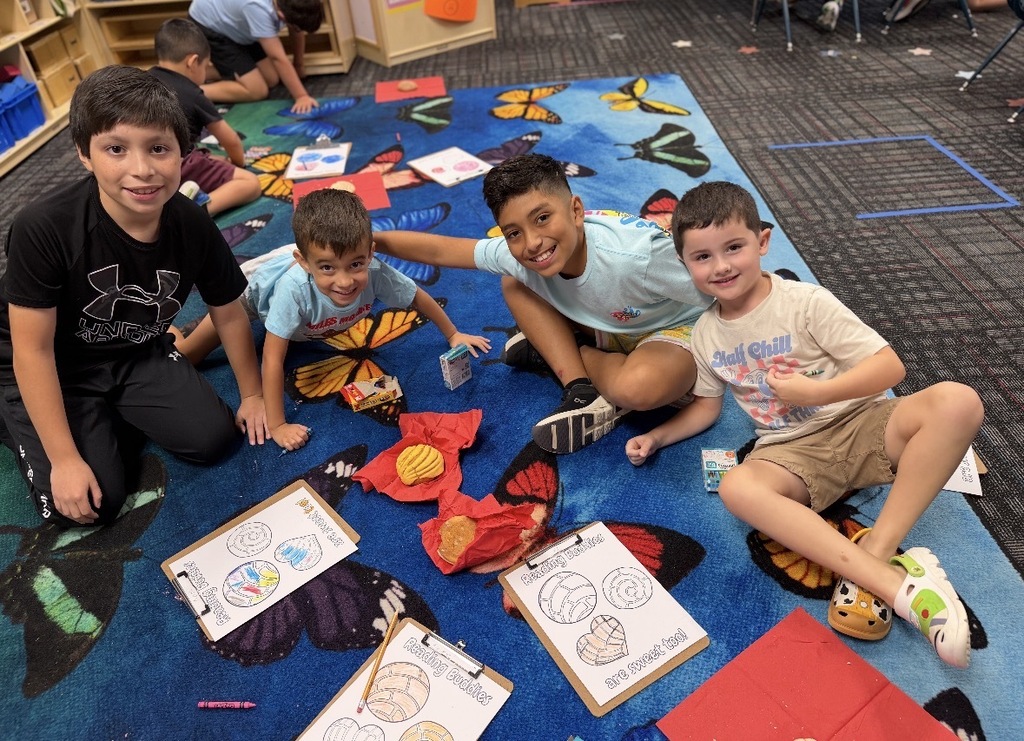 Four young students smile together while sitting on a colorful butterfly-patterned classroom rug. They are surrounded by "Reading Buddies" clipboards with coloring pages, crayons, and pieces of sweet bread (conchas) resting on red napkins.