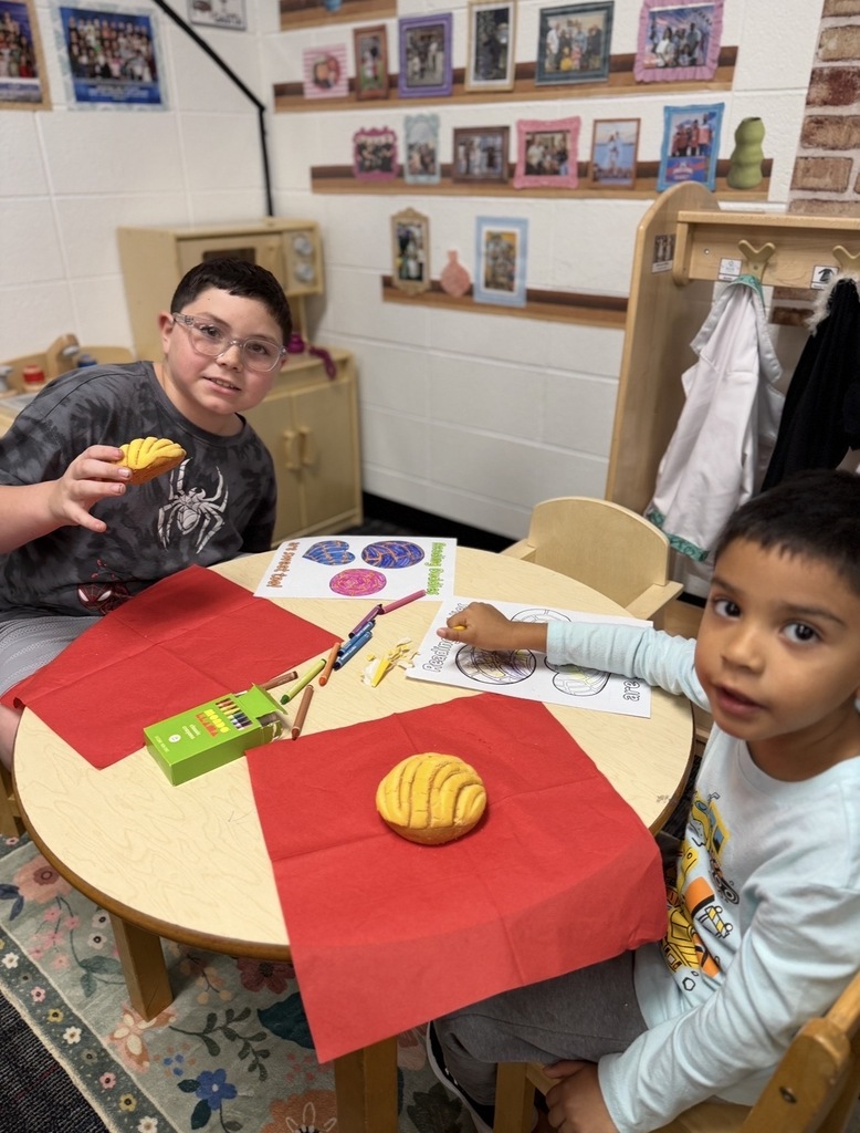 Two young boys sit at a small round wooden table during a "Reading Buddies" activity. The boy on the left, wearing glasses and a gray spider-graphic shirt, holds up a piece of sweet bread (concha), while another concha rests on a red paper napkin in front of the younger boy on the right. Between them on the table are crayons, red napkins, and "Reading Buddies are sweet too!" coloring sheets featuring pan dulce illustrations.