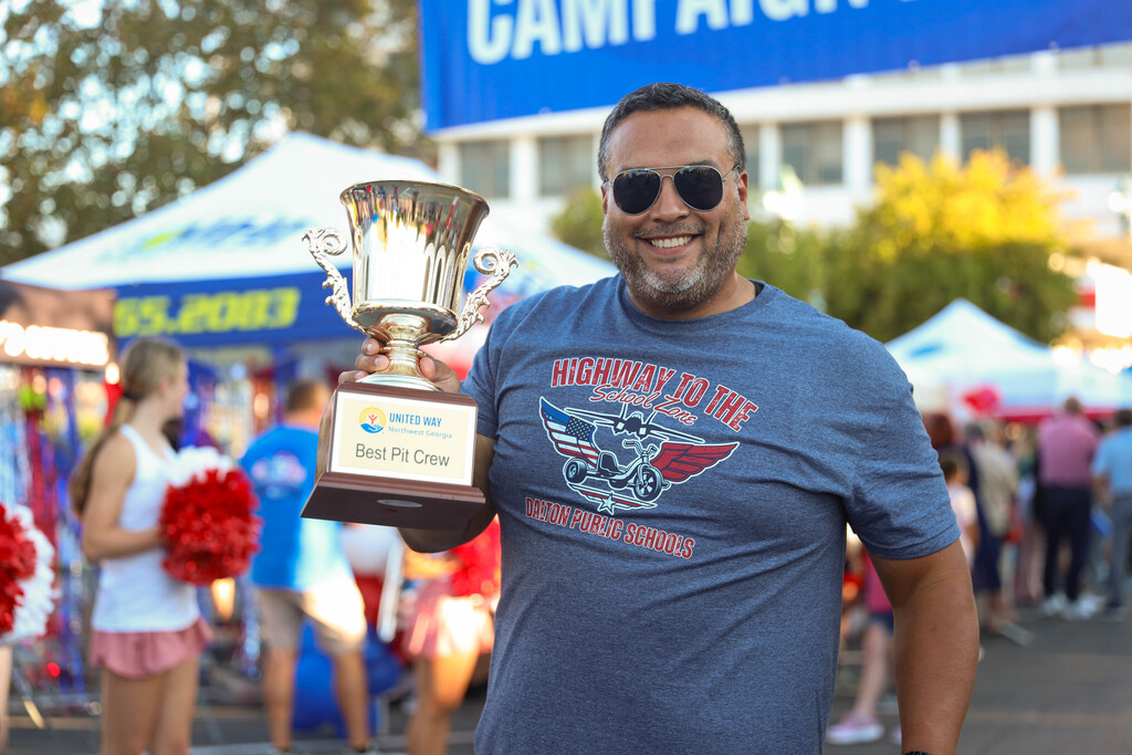 A school staff member holding up the United Way "Best Pit Crew" award trophy outdoors.