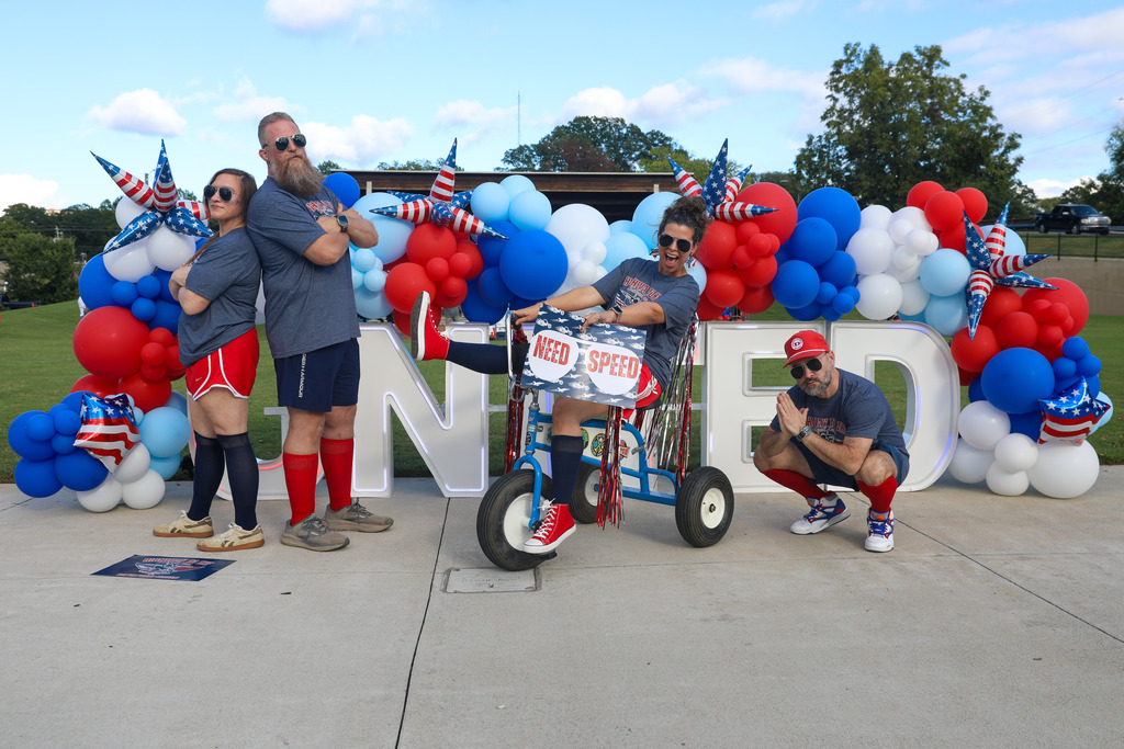 A pit crew team posing with a tricycle in front of a red, white, and blue balloon arch.
