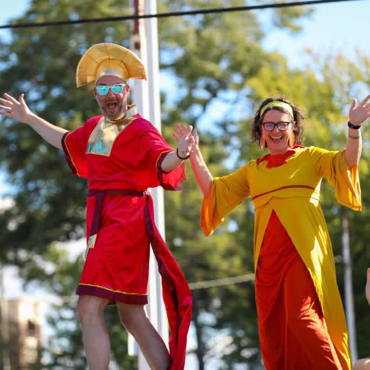 Two people in vibrant red and yellow costumes smile and wave enthusiastically while riding on a parade float under a bright, sunny sky.