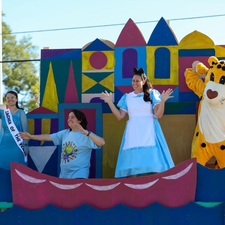Parade float participants costumed as a teacher of the year, a person in a blue dress, and a cheetah mascot stand on a colorful "It's a Small World" themed float waving to the crowd.