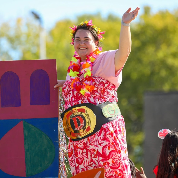A smiling woman wearing a floral dress, lei, and a championship belt waves to spectators from a parade float decorated with colorful geometric shapes