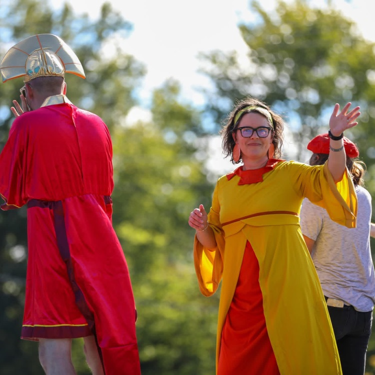Parade participants in vibrant red, yellow, and traditional costumes wave and interact with spectators from the side of a parade float.