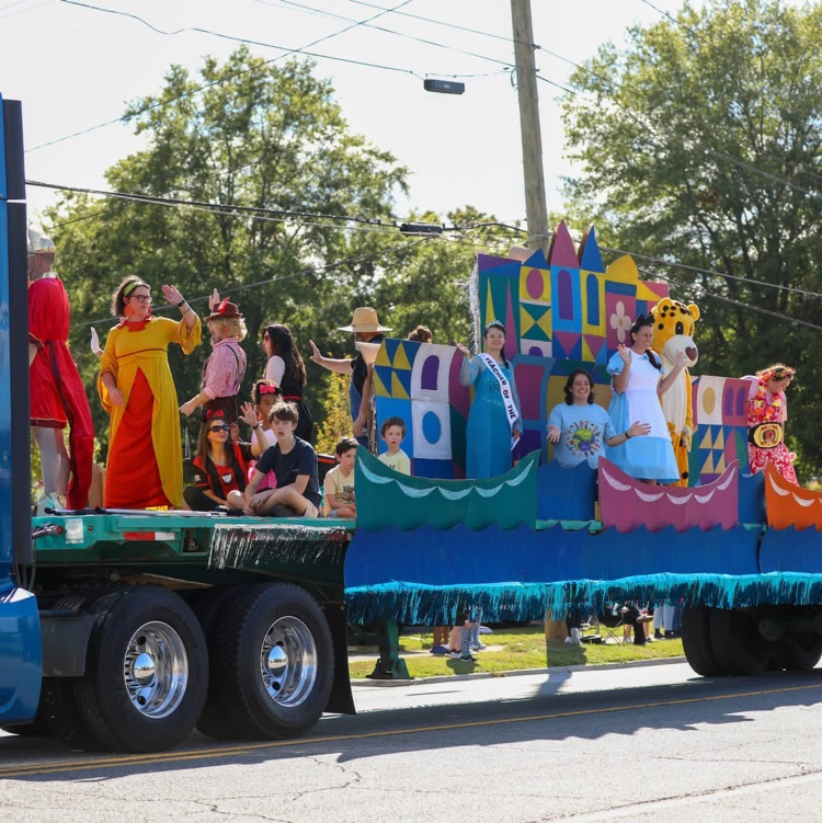 A large blue semi-truck pulls a long flatbed parade float filled with costumed adults and children waving to onlookers along a tree-lined stree