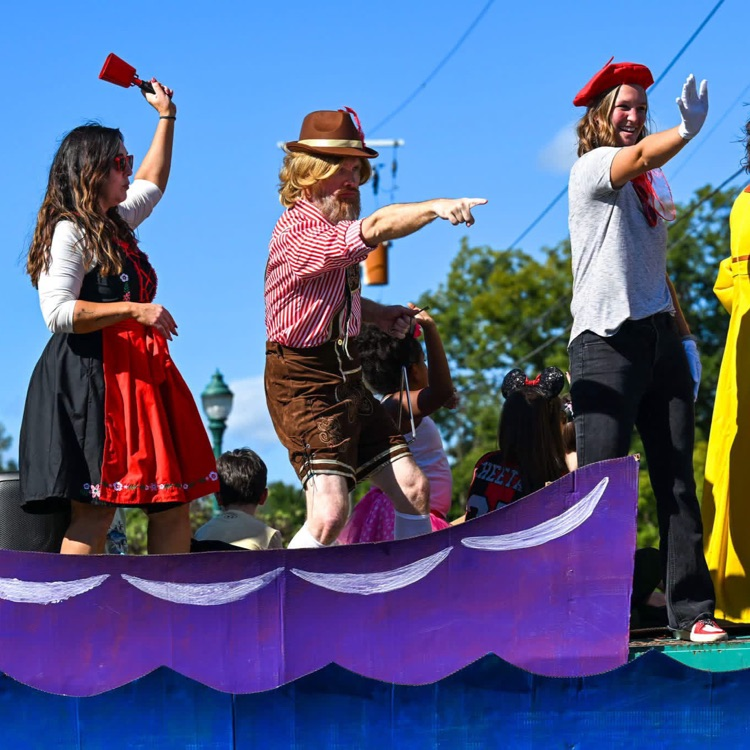 Four costumed parade participants stand atop a float, including two wearing lederhosen and traditional attire, waving and pointing toward the crowd