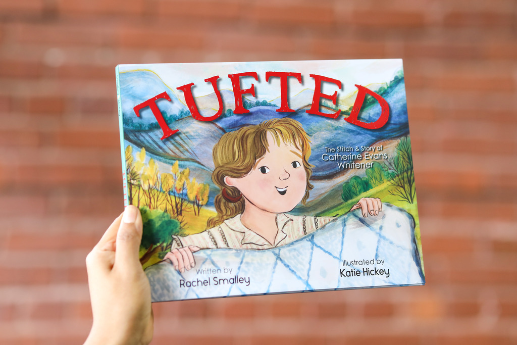 A hand holding up a copy of the children's book Tufted: The Stitch & Story of Catherine Evans Whitener against a brick wall background.