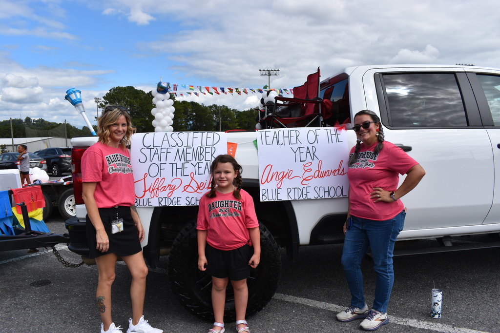 Main Subject: Two women and a young girl standing in front of the side bed of a white pickup truck, posing for a photo.   People & Attire:On the left, a woman (Tiffany Seil) in a pink Blue Ridge shirt and black skirt smiles next to a poster.   In the center, a young girl in a matching pink shirt reading "DAUGHTER" and black shorts stands proudly.   On the right, a woman (Angie Edwards) wearing a pink Blue Ridge shirt, blue jeans, and sunglasses poses with her hand on her hip.   Signs & Recognition:Left poster: "CLASSIFIED STAFF MEMBER OF THE YEAR Tiffany Seil - BLUE RIDGE SCHOOL".   Right poster: "TEACHER OF THE YEAR Angie Edwards - BLUE RIDGE SCHOOL".   Setting: An outdoor parking lot setup for a parade or event on a partly cloudy day, with a trailer attached to the pickup truck. 