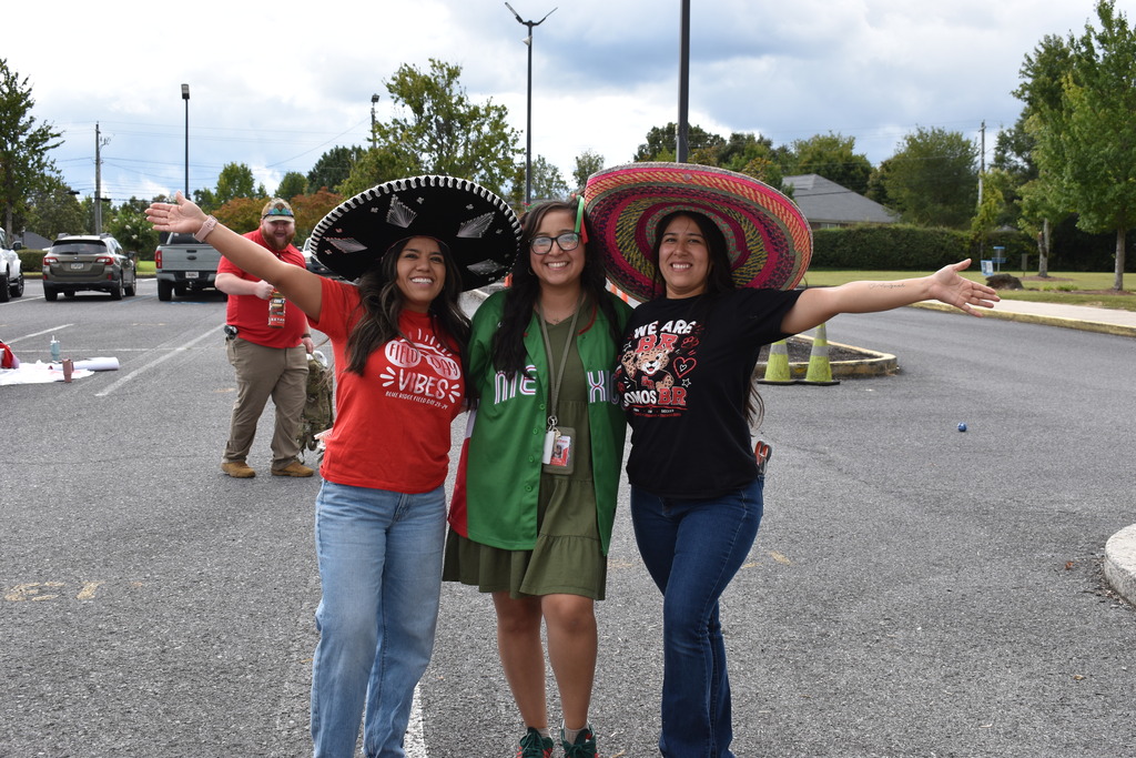 Main Subject: Three women pose closely together in an open outdoor parking lot with wide smiles, arms outstretched to the sides.   Attire & Accessories:The woman on the left wears a large black and silver embroidered sombrero, a red t-shirt, and blue jeans.   The woman in the middle wears glasses, a green dress layered with an open green Mexico jersey, and an ID badge lanyard.   The woman on the right wears a wide-brimmed colorful pink and yellow sombrero, a black "WE ARE BR" t-shirt, and dark jeans.   Background: In the background, a man in a red shirt and khaki pants stands near parked cars in an asphalt parking lot under a cloudy sky.
