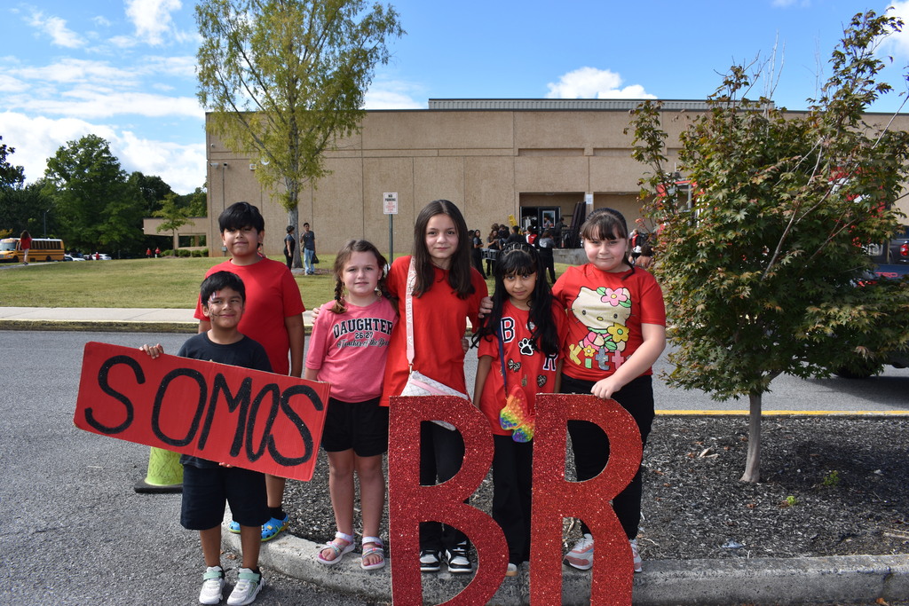 Main Subject: A group of six elementary students standing together outside near a sidewalk curb posing for a picture.   Props & Signs:A young boy on the left holds a long red cardboard sign with black lettering that reads "SOMOS".   Standing in front of the middle and right students are two large, standalone red glitter cutout letters forming "BR".   Clothing: Most students are wearing red shirts, with one girl in a pink shirt reading "DAUGHTER" and another wearing a Hello Kitty top.   Background: The photo is taken outside near a parking lot in front of a multi-story school building with trees, grassy hills, and a yellow school bus visible under a partly cloudy sky.  