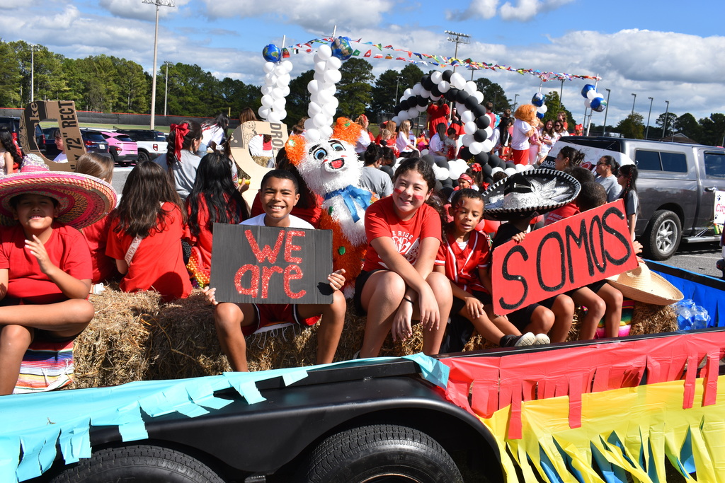 Main Subject: A group of cheerful elementary students sitting on hay bales aboard a decorated trailer float during a parade.   Signs & Props:A smiling boy in the center holds a black rectangular sign with glittery red text that reads "WE ARE".   A student next to him holds a red sign with black lettering that reads "SOMOS".   Several children are wearing large sombreros, including a black and silver embroidered one and a wide-brimmed pink one.   Float Decorations:A white bunny/mascot piñata with an orange wig and blue bow sits among the students.   The sides of the trailer are decorated with blue, red, and yellow fringe and streamers.   Black and white balloon arches span the background of the trailer float.   Setting: Outdoors on a clear, sunny day with bright blue skies, light clouds, and trees visible behind parked vehicles along the parade route.   