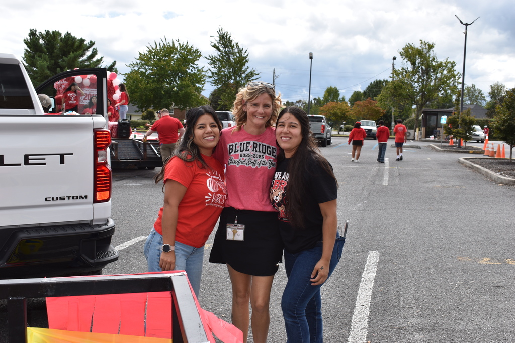 Main Subject: Three smiling women stand arm-in-arm posing for a photo in an outdoor parking lot area.   Clothing & Details:The woman on the left wears a red t-shirt and light blue jeans.   The woman in the center wears a pink shirt reading "BLUE RIDGE 2025-2026 Classified Staff of the Year" with a black skirt and a lanyard badge.   The woman on the right wears a black "WE ARE BR" shirt and blue jeans.   Setting & Background: They are standing next to the bed of a white Chevrolet pickup truck. In the background, other participants in red shirts walk through a asphalt parking lot, and a trailer decorated with red balloons for a parade float is visible under an overcast sky.   