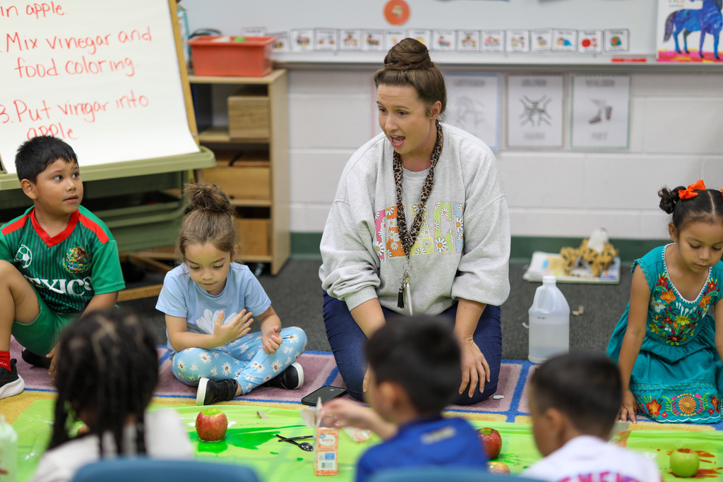 A teacher speaks to student sitting on a rug.