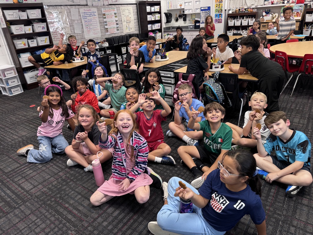 A wide shot of a elementary classroom filled with smiling students enjoying a snack activity. A large group of children sit on the floor in the foreground, many holding up small round cookies toward the camera, while other classmates sit and pose at tables throughout the room.
