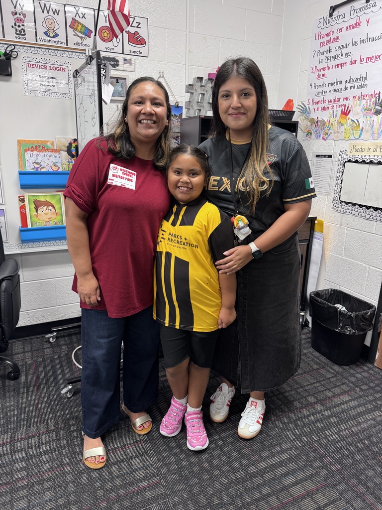 A young girl in a yellow-and-black sports jersey stands proudly between two smiling women inside a classroom. The woman on the left wears a red shirt with a visitor badge, while the woman on the right wears a dark shirt. Classroom charts, alphabet cards, and Spanish learning materials line the wall behind them.