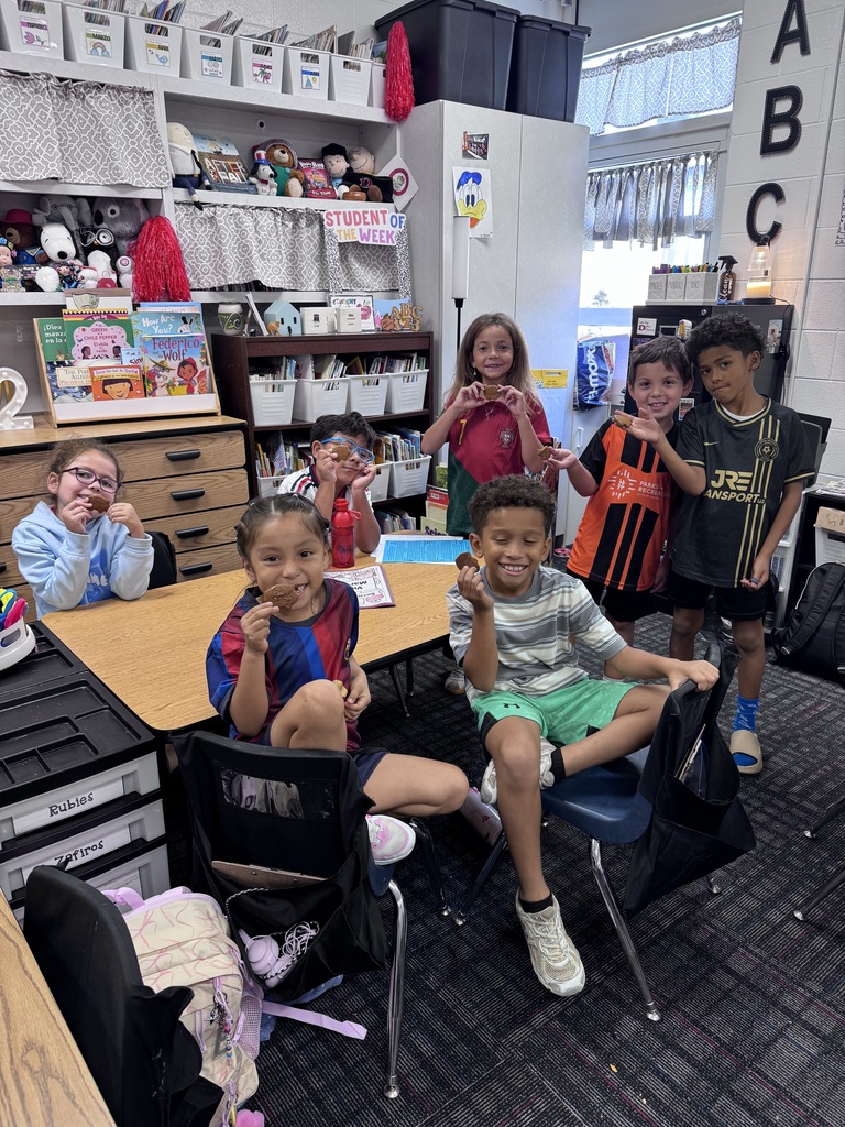 Six young students gather around a low table in a organized classroom, smiling as they show off small brown cookies. Two students sit in the front foreground, one in a striped shirt and another in a soccer jersey, while four classmates smile and pose behind them near bookshelf storage bins and a "Student of the Week" display board.