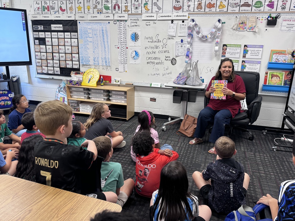A teacher sits at the front of a dual-language classroom, holding up a colorful children's book to read aloud to a group of elementary students sitting on the carpet. The classroom walls are filled with educational posters in Spanish and English, calendar charts, and alphabet banners.