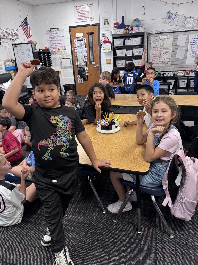 An elementary school student wearing a colorful dinosaur shirt stands in the foreground, smiling as he holds up a small cookie above his head. Behind him, several classmates sit at a wooden desk enjoying their cookies, surrounded by classroom educational posters and supplies.