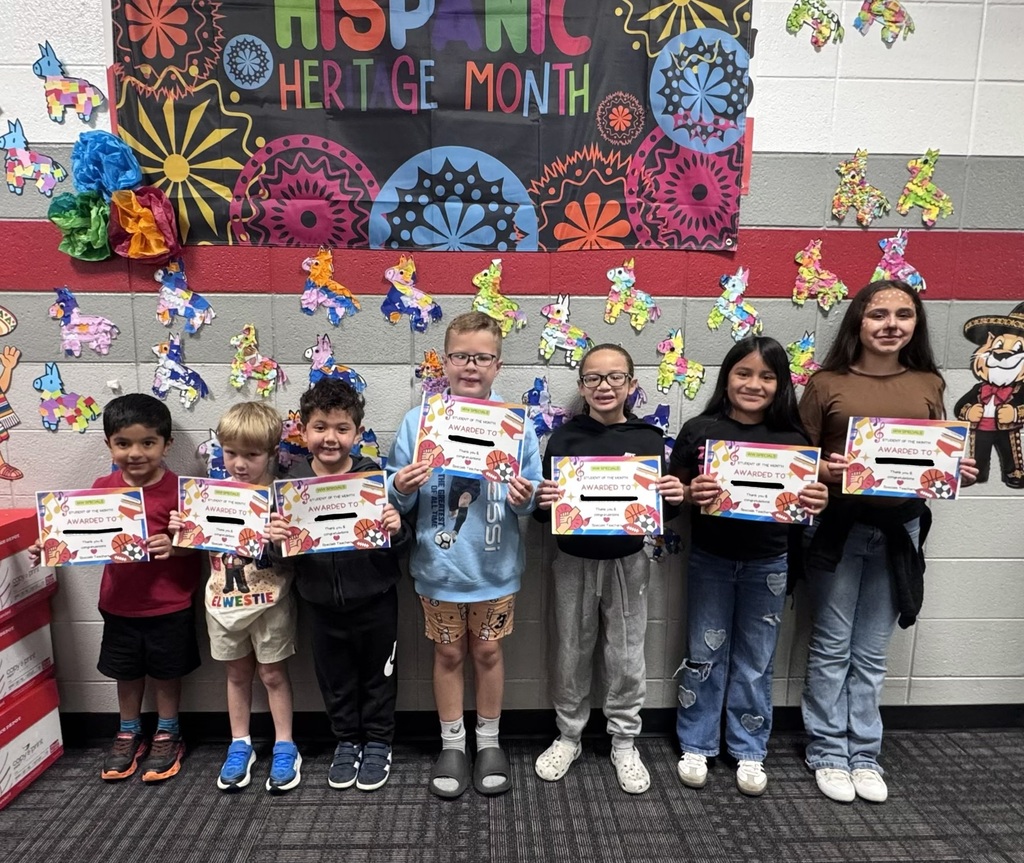 Seven elementary school students stand side-by-side in a school hallway holding "Student of the Month" certificates. Behind them, the wall is decorated for Hispanic Heritage Month with a black banner featuring colorful text and patterns, along with several small, multi-colored piñata cutouts scattered across the wall.