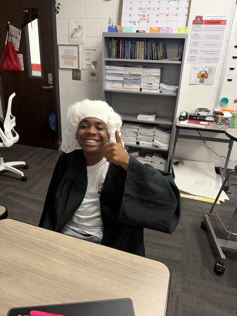 A student wearing a judge’s robe sits at the front of the classroom during a mock trial activity, listening to classmates present evidence and defend their arguments.