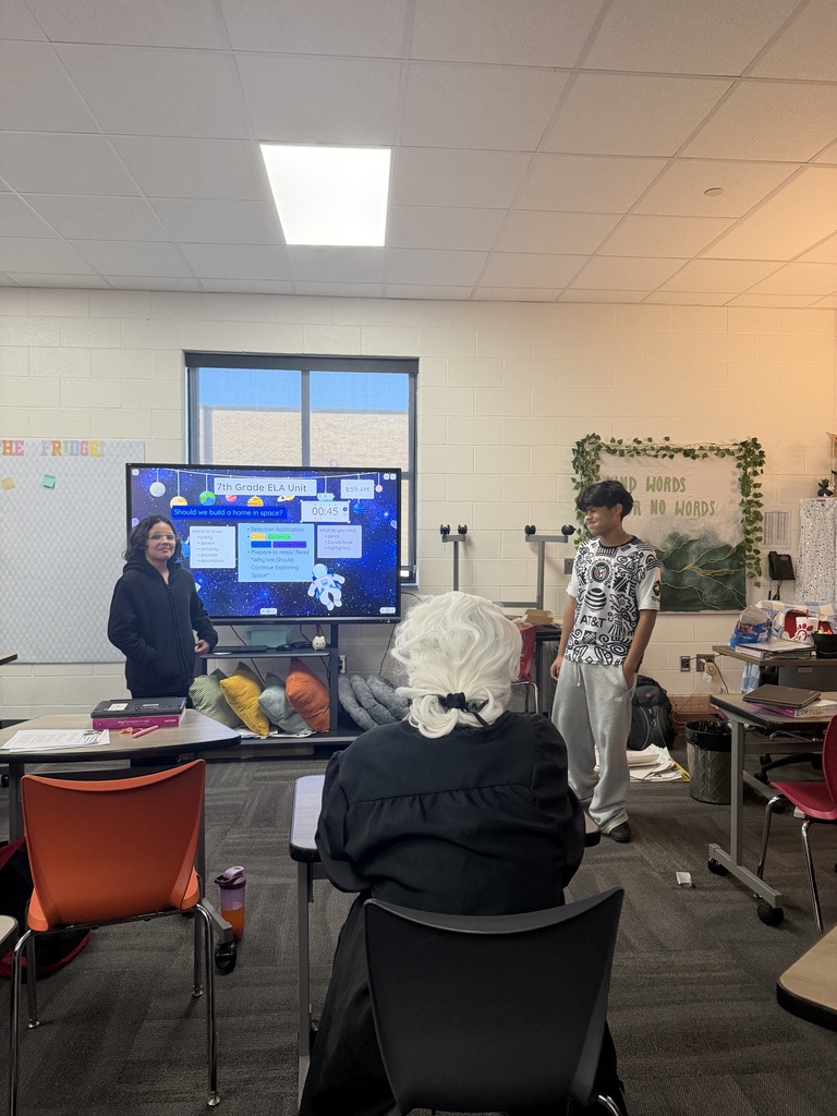 Two students stand at the front of the classroom presenting their case during a mock courtroom activity, using evidence and reasoning to support their argument.